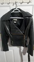 Black women's leather jacket hanging on a white hanger, showing front with zipped pockets and asymmetrical zipper.