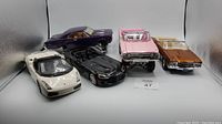 Five metal model cars on display showing variety in colors and styles, including a purple muscle car, pink convertible, black and white sports cars, and a bronze coupe.