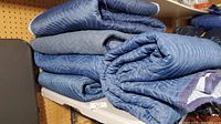 Stack of five blue moving blankets folded and stacked on a shelf, showing the texture and thickness of the fabric.