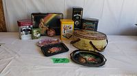 Photo showing collection of vintage tins and hand-painted metal trays on a white tablecloth against a white paneled background. Includes several decorative tins and an oval carrier with leather handles.