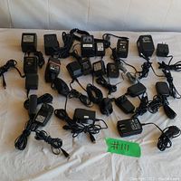 Wide top-down photo showing 23 black AC power adapters for cellphones and other electronic devices laid out on white surface.