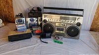 Overview of lot showing large vintage boombox, smaller speaker, handheld radios, digital radio alarm clock, and sealed cassette tapes in front