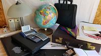 Photo showing vintage world globe, two briefcases, calculators, binders, desk lamps, pens, and accessories on a table