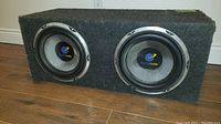 Dual 12 inch Planet Audio subwoofers installed in a rectangular carpeted wooden enclosure, front view showing subwoofers with visible repair on one.