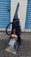 Full front view of the upright carpet cleaner showing the handle, hose, and transparent cleaning head with labels.
