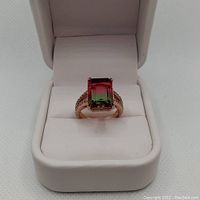 Top front view of rose gold plated ladies ring with a large rectangular bi-color green to red stone in claw setting inside a white ring box.