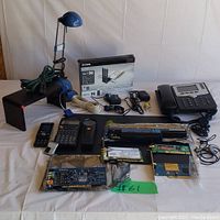 Full lot overview showing all items spread on white surface including computer boards, phone accessories, office phone, calculator, and lamp.