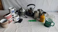 Full lot view with electric egg cooker, kettle, ceramic mugs, metal mugs, glass mugs and yellow juicer arranged on table