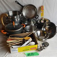Full view of the lot showing pots, pans, strainers, and box of utensils and plastic wrap on a white cloth background.