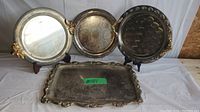 Four serving trays displayed on stands and flat, showing tarnish, wear, and scratches on silver plate and metal surfaces.