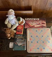 Photo shows Scrabble board, two Scrabble books, Ginnykub board game box, and three stuffed animals including a rabbit, a duck, and a bear on a carpeted floor.