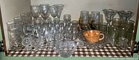 Wide view of entire lot, showing multiple types of cut glass aperitif glasses, amber glass mugs, parfait glasses, bowls and stemware arranged on checked cloth.