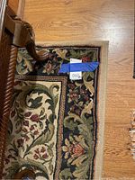Photo showing natural hardwood floor and corner of a table leg on the edge of a beige, black, burgundy floral patterned area rug with visible price tag.