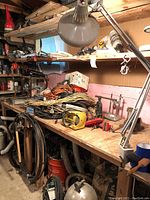 Workshop bench with multiple coils of electrical cables, an adjustable desk lamp, various hand tools, and hardware items scattered on the wooden work surface and shelves.