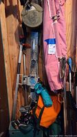 Wide shot of skis, ski poles, walking sticks, orange backpack, pink camping chair bag, and small round green pouch hanging on wooden wall.