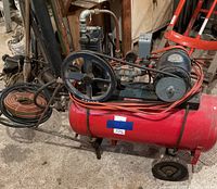 Red horizontal air compressor tank with motor and belt. Two hoses coiled beside compressor. Hand truck frame visible.