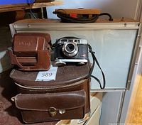 Kodak film camera with lens, showing body and strap, accompanied by two brown leather cases, one larger carrying case and one smaller pouch, displayed on a surface in front of a backlight box.