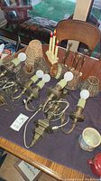 Table displaying several vintage brass wall lamps with bulbs, various woven and pleated lampshades, two candle stick holders, and ceramic coffee cups.