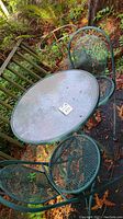 Metal outdoor table with clear plastic top and four green metal foldable mesh chairs around it, set on outdoor ground with leaves.