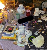 Wide shot showing miscellaneous household items including ceramics, glassware, mugs, pins, fabric doilies, and a small lamp on table