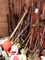 Photo showing an assortment of outdoor tools including pitchforks, shovels, and long handled tools leaning against a wall with wooden background, plus red gas cans on the floor.