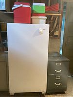 Full view of white Kenmore upright freezer with four assorted coolers on top and gray filing cabinet to the side.