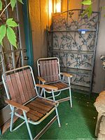 Two metal framed folding chairs with wooden slats and armrests in front of metal shelving unit with decorative vine and grape design