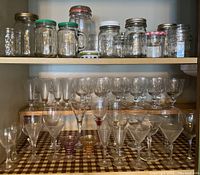 Shelf with clear glass cut goblets in back, assorted martini glasses and aperitif glasses in front, top shelf with various glass canning jars with metal lids.
