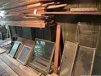 Photo showing multiple vintage wooden window frames lined on floor underneath stacked wood trim above, highlighting variety of sizes and condition.