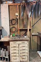 Overall view of wooden workbench with drawers underneath, hand saws mounted above, assorted tools and wood dowels on shelf and workbench surface.