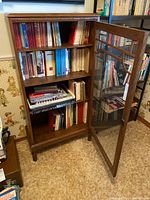 Front angle of wood cabinet with glass door open revealing books on shelves inside.