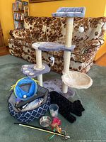 Wide shot showing cat tree with three levels, paw-shaped platforms, sisal scratching posts, and base. Various cat care accessories including cat bed, tunnel, toys, and feeding bowls arranged on floor in front.