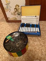 Boosey & Hawkes student glockenspiel in blue plastic case with yellow foam lining, wood mallets, and steel drum with multicolor striped body and hand-drawn design.