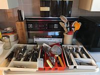 Two trays of kitchen cutlery, utensils, and accessories arranged on a stove including cutlery block, spoons, forks, knives, and kitchen gadgets.