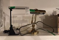 Photo of two desk lamps, one black adjustable OTT Lite, one brass with green glass shade, and three metal sock stretcher frames on floor.
