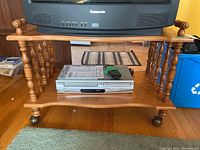 Front view of the solid wood media cart holding a Panasonic CRT TV and a DVD player on the lower shelf, showing the turned spindle side rails and caster wheels.