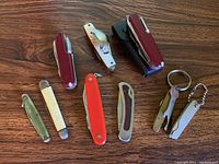 Nine assorted pocket cutlery pieces on wooden surface, showing various colors and styles including the black protective case and keychain knives.