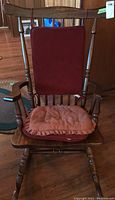 Front view of the wooden rocking chair showing reddish cushions on seat and backrest, spindle detailing on back and turned legs.
