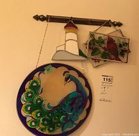 Five hanging stained glass suncatchers including a round peacock, rectangular floral floral panel, cardinal bird panel, lighthouse design, all hanging on a decorative metal hook on the wall.