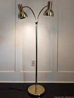 Full view of brass floor lamp showing round weighted base, vertical pole, and two adjustable lamp heads facing outward.