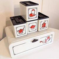 Front and angled view of the vintage metal breadbox with four matching metal kitchen canisters arranged on top, showing their red illustrations and black lids.