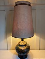 Side view of the lamp turned on, showing the shape of the beige fabric shade with brown accent stripes and the mottled multicolor ceramic base reflecting light.
