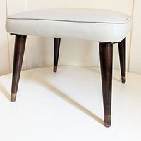 Front angled view of footstool showing light gray vinyl padded seat and tapered dark wooden legs with metal feet caps.