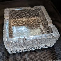 Close-up angled view showing the textured surface and cigarette rest notch on the square glass ashtray.