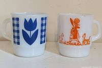 Front view of two mugs side by side showing their front prints, blue tulip and orange girl with dogs.