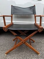 Front view of vintage folding directors chair showing wooden frame, black fabric seat and backrest, wooden armrests, cross-braced legs on an asphalt surface.