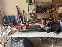 Photo of the yard tools including the leaf blower, chainsaw, and weed eater arranged on a white folding table with garage shelving in the background.