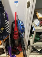 Photo shows Shark and Dirt Devil upright vacuums together with a Swiffer mop in a closet space.