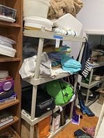 Gray plastic shelving unit with folded beach towels, blankets, paper plates, plastic bowls, and electric grill stored on shelves.