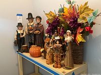 Wide view of the fall decor pieces on top of a white shelf against a gray wall showing pilgrim family figures and floral arrangements.
